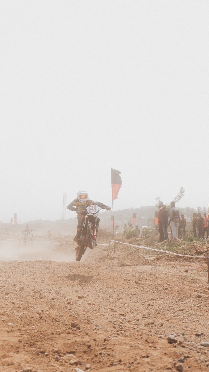 Offerings A motocross rider performing a jump on a dirt track amidst a dusty environment with spectators.